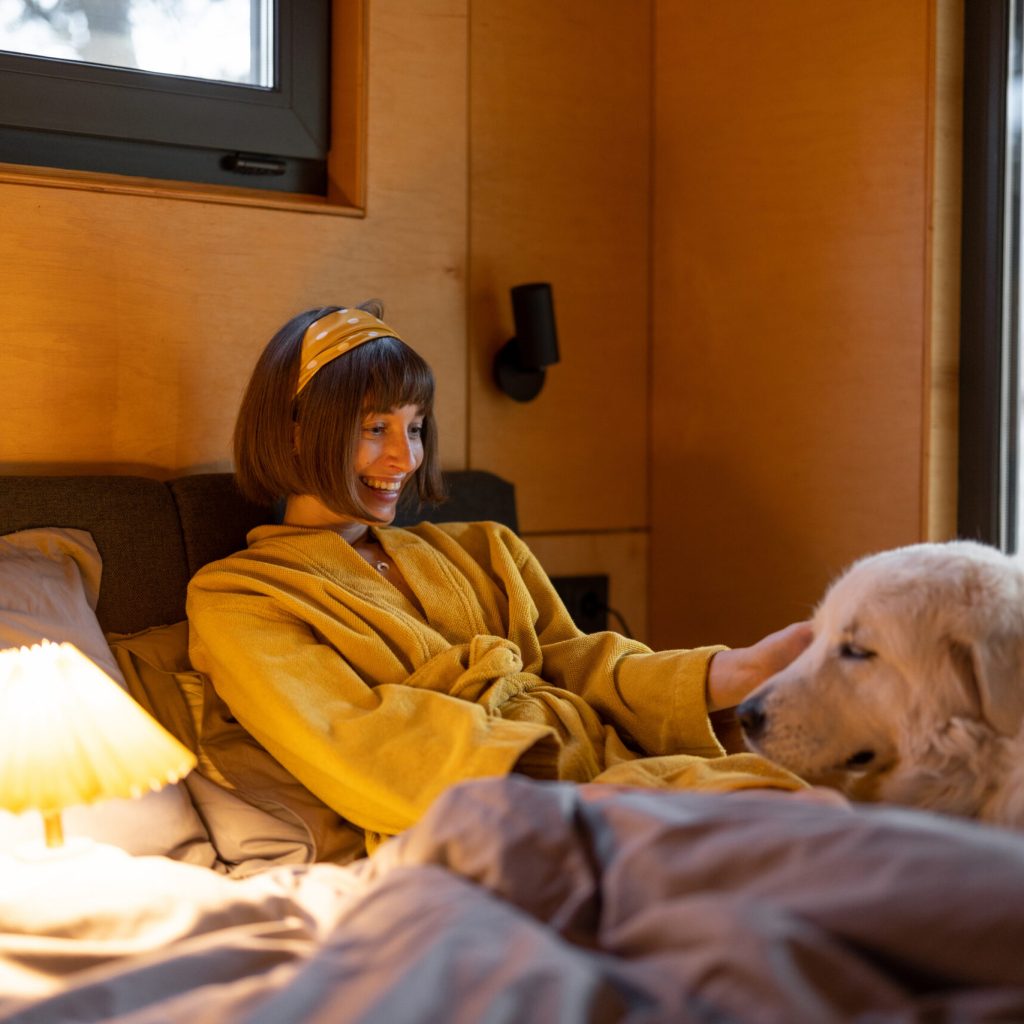 Young woman cares her huge adorable white dog while lying in bed in tiny bedroom of wooden cabin on nature. House coziness and friendship with pets concept